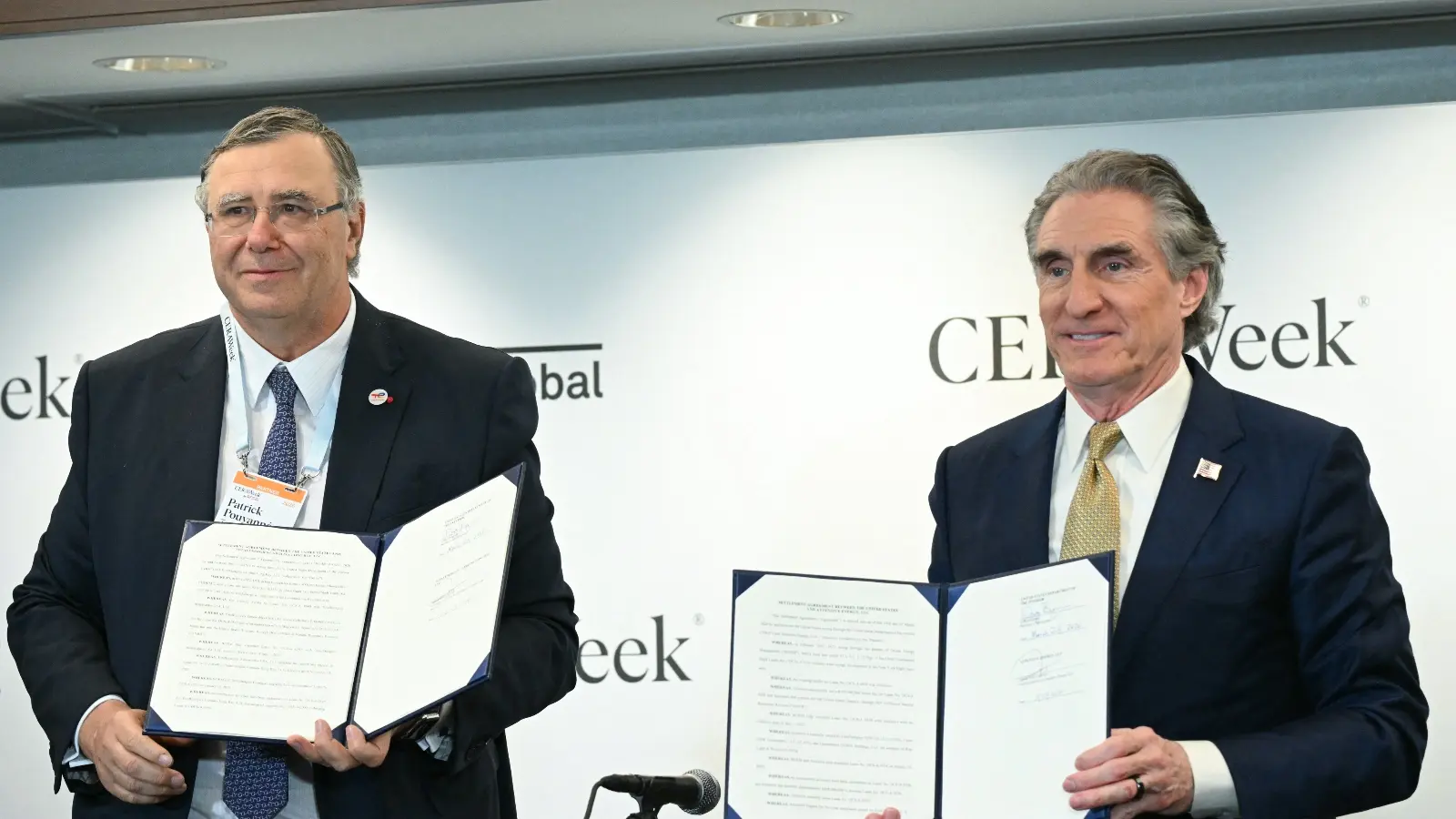 Two men in business suits stand in front of a white background. They are holding papers.