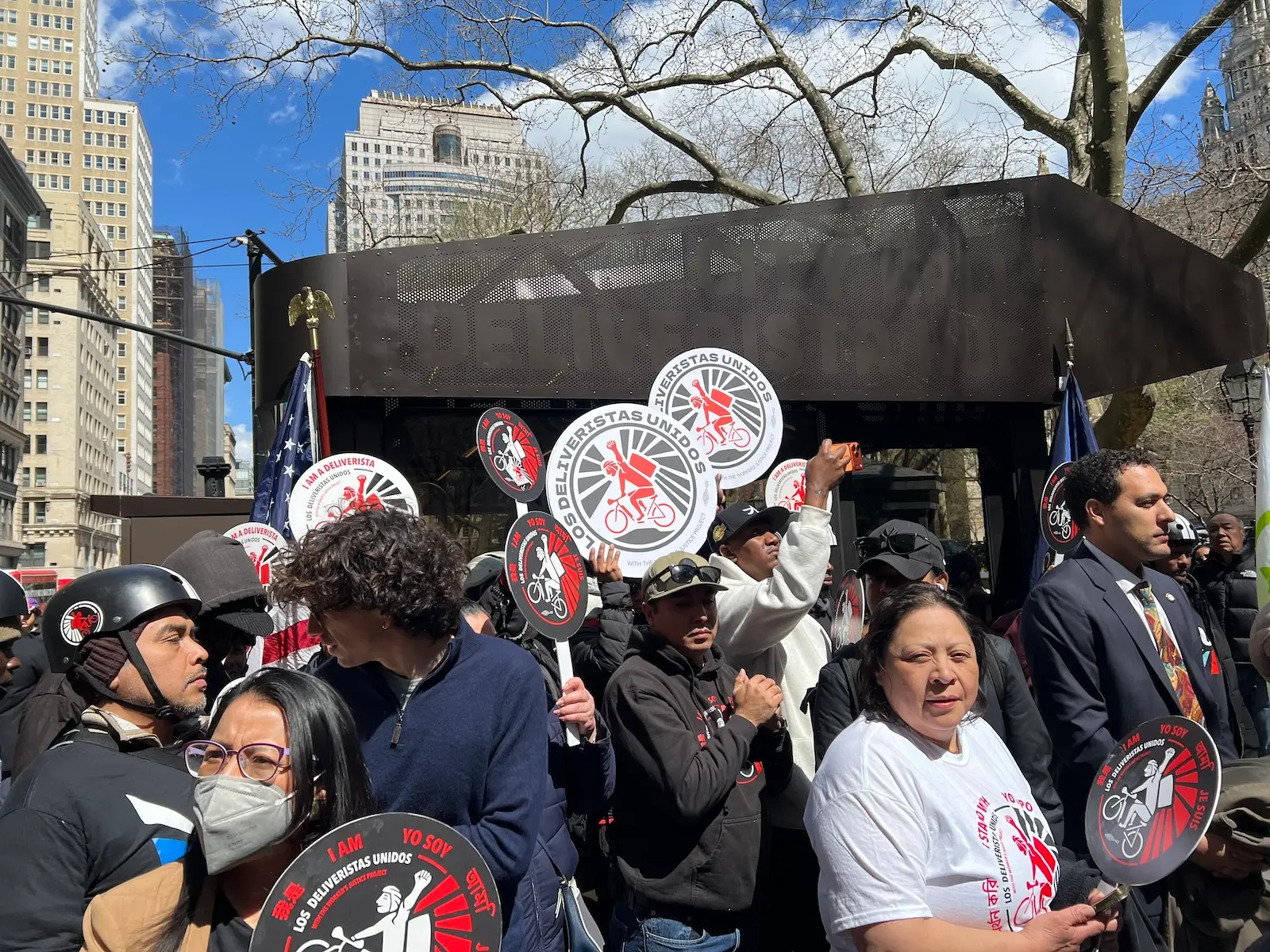 A group of app-based delivery workers hold up signs for their union, Los Deliveristas Unidos, while standing next to New York City's first-ever rest stop for delivery workers. The hub is an industrial-looking, metallic structure. There is a tree and a blue sky with some clouds in the background, and some buildings to the left.