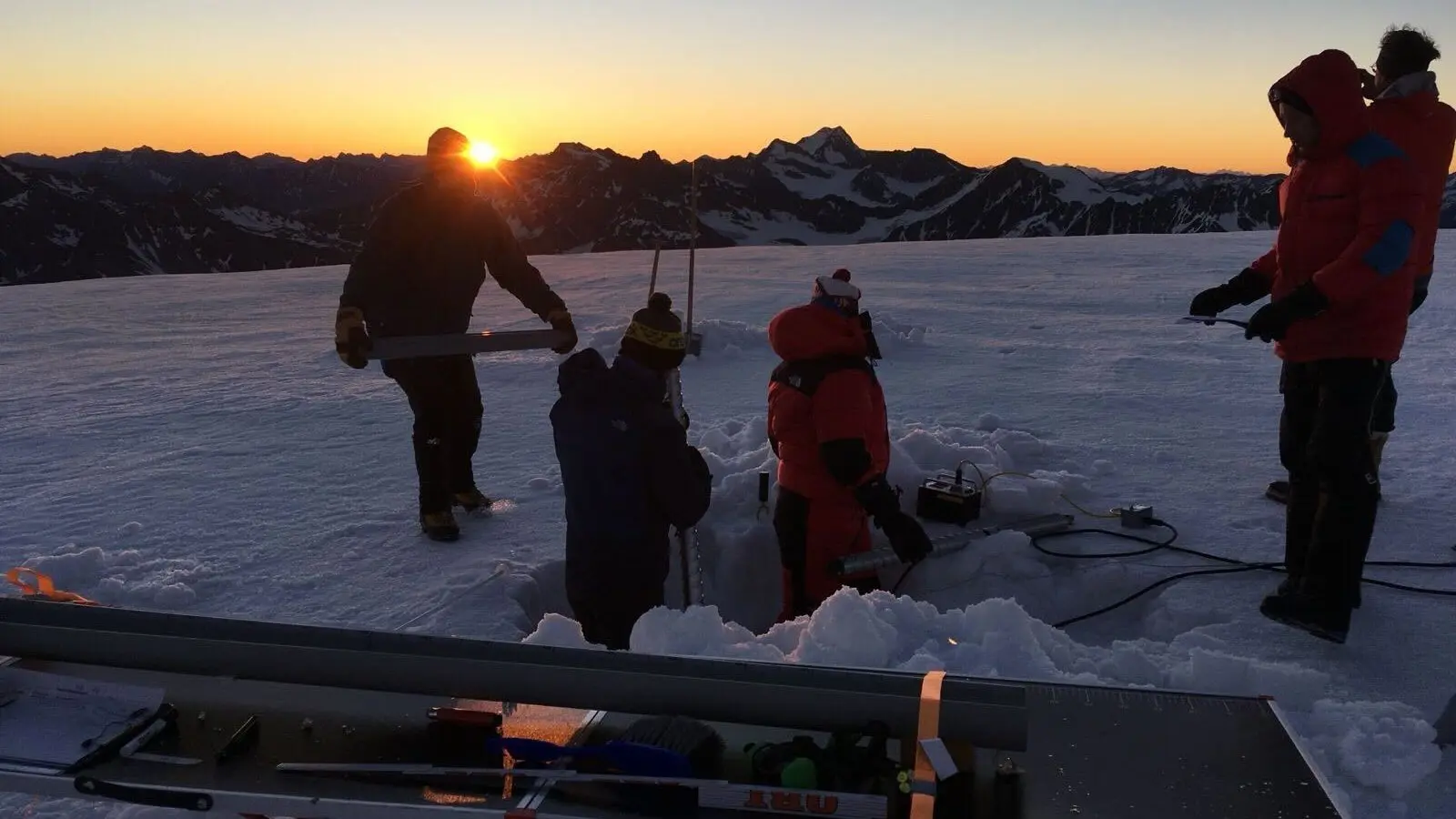 Four researchers in coats and hats stand around a hole dug in the ice on top of a glacier, while the sun peeks over the mountains behind them
