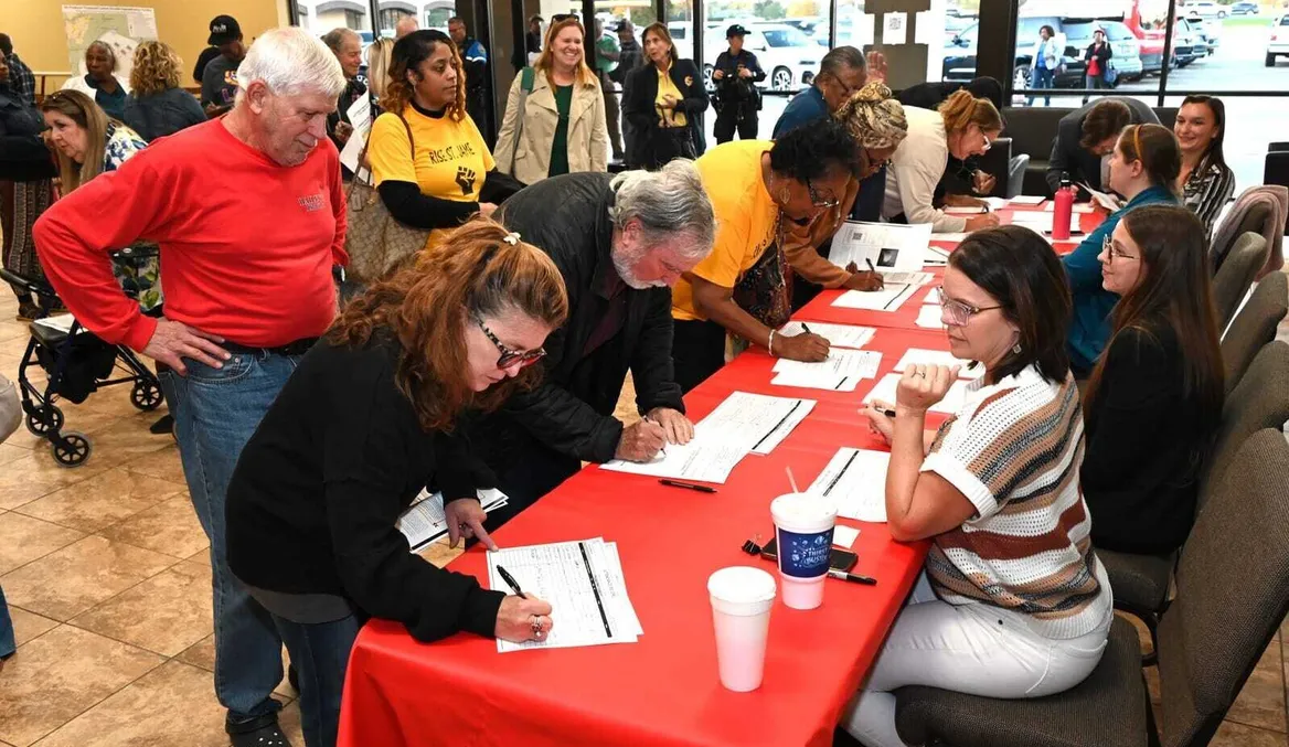 A large group of people in a room, some signing papers on red tablecloth with four people sitting behind the table