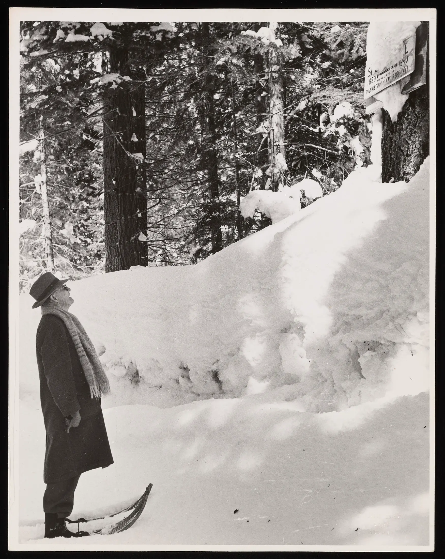 A man in a black and white photo stares up at a snow drift. He is wearing skis and a coat and hat