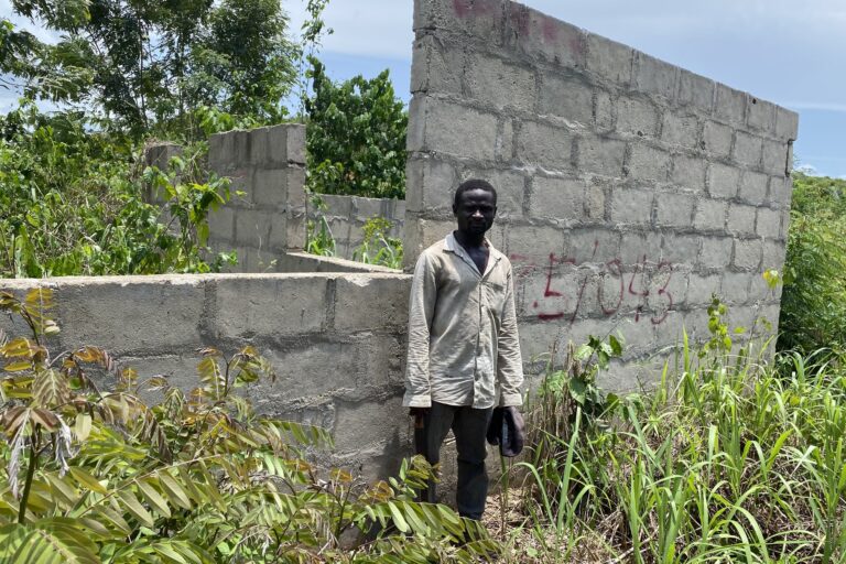 Samuel Nkrumah stands next to his unfinished two-bedroom house, which has been earmarked for demolition. He currently lives with his parents and will be resettled with the rest of the village of Krampakrom. Image by Maxine Betteridge-Moes for Mongabay.