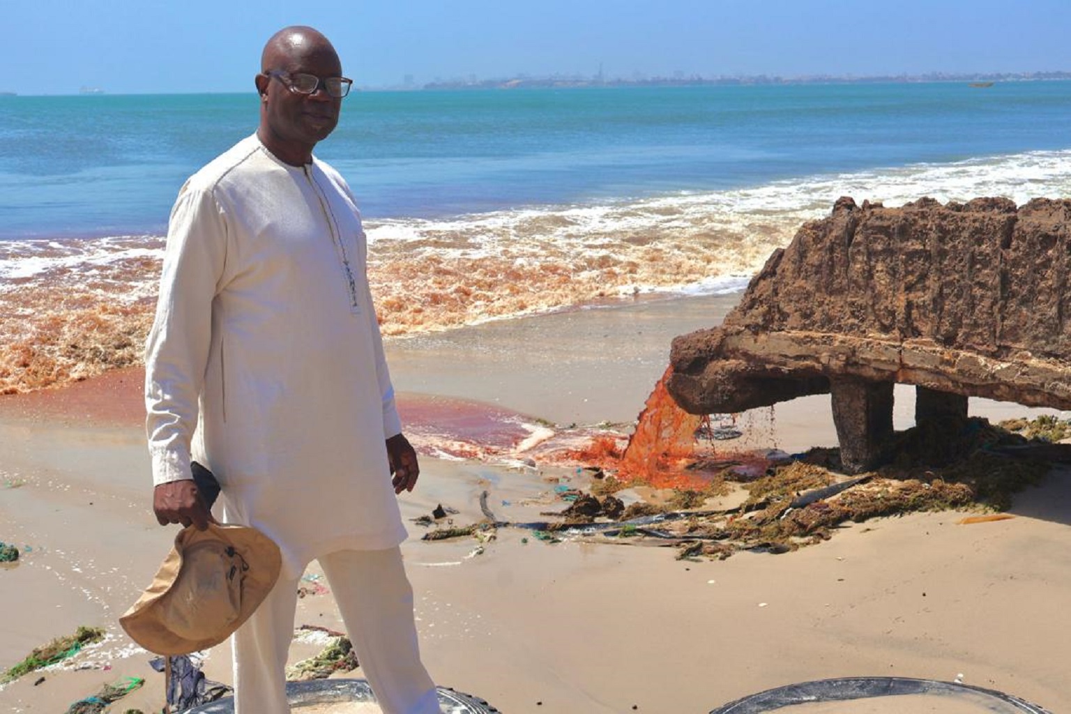 Mbacke Seck, executive director of Hann Baykeeper, a coastal community organization in Senegal, photographed alongside Hann Bay in March 2026. Some businesses there discharge their waste into the bay , the pipe shown is connected to a slaughterhouse company, according to Seck. The city of Dakar, Senegal’s capital, can be seen in the distance.