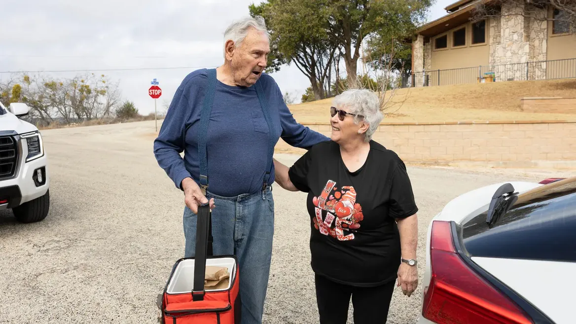 Woman with white hair and glasses on the side of a road next to a man in jeans and a blue top, holding a red cooler
