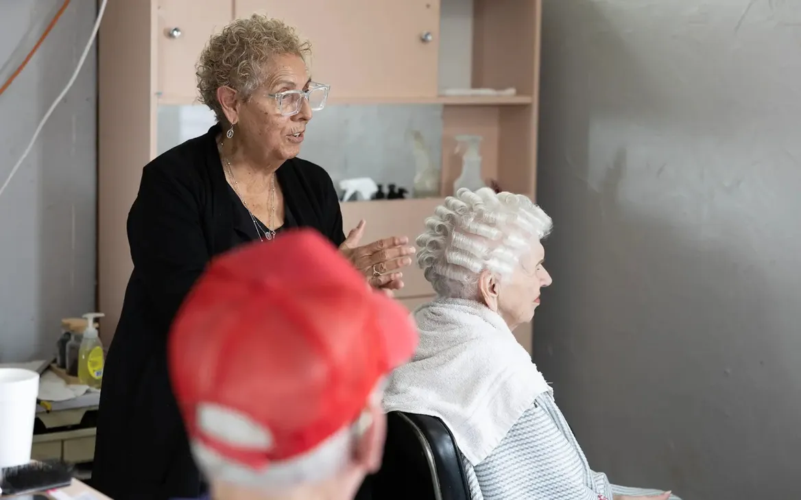 A hair stylist works on a woman whose hair has been in rollers , a man in a red cap watches.
