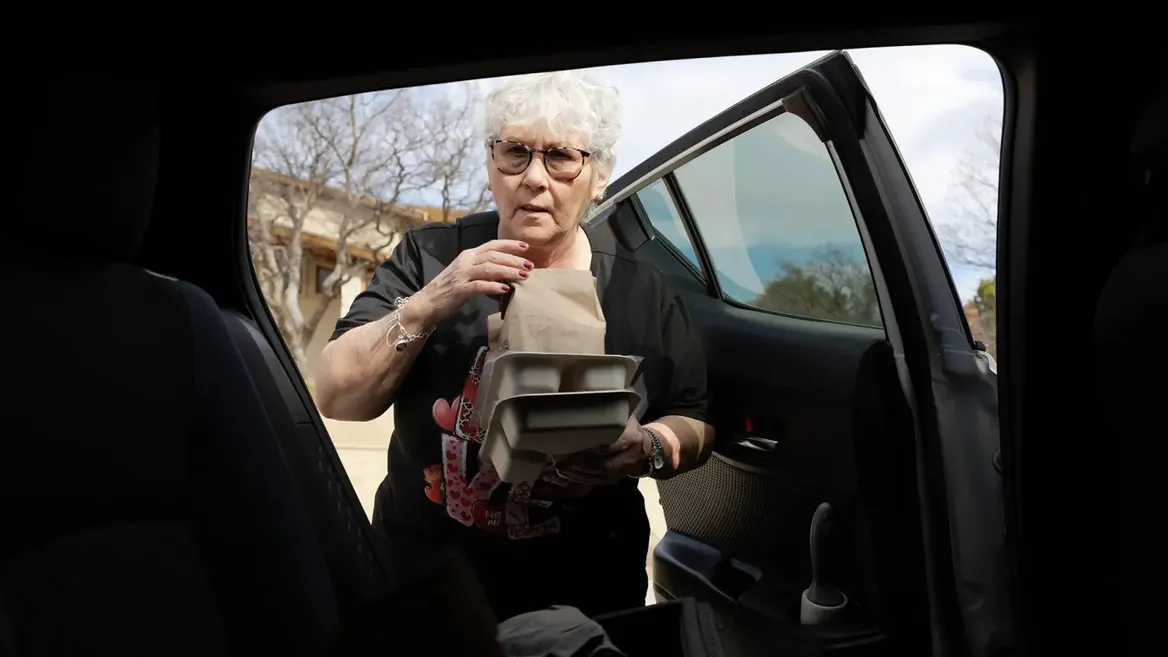 A woman with glasses and white standing in front of an open car door holding food containers