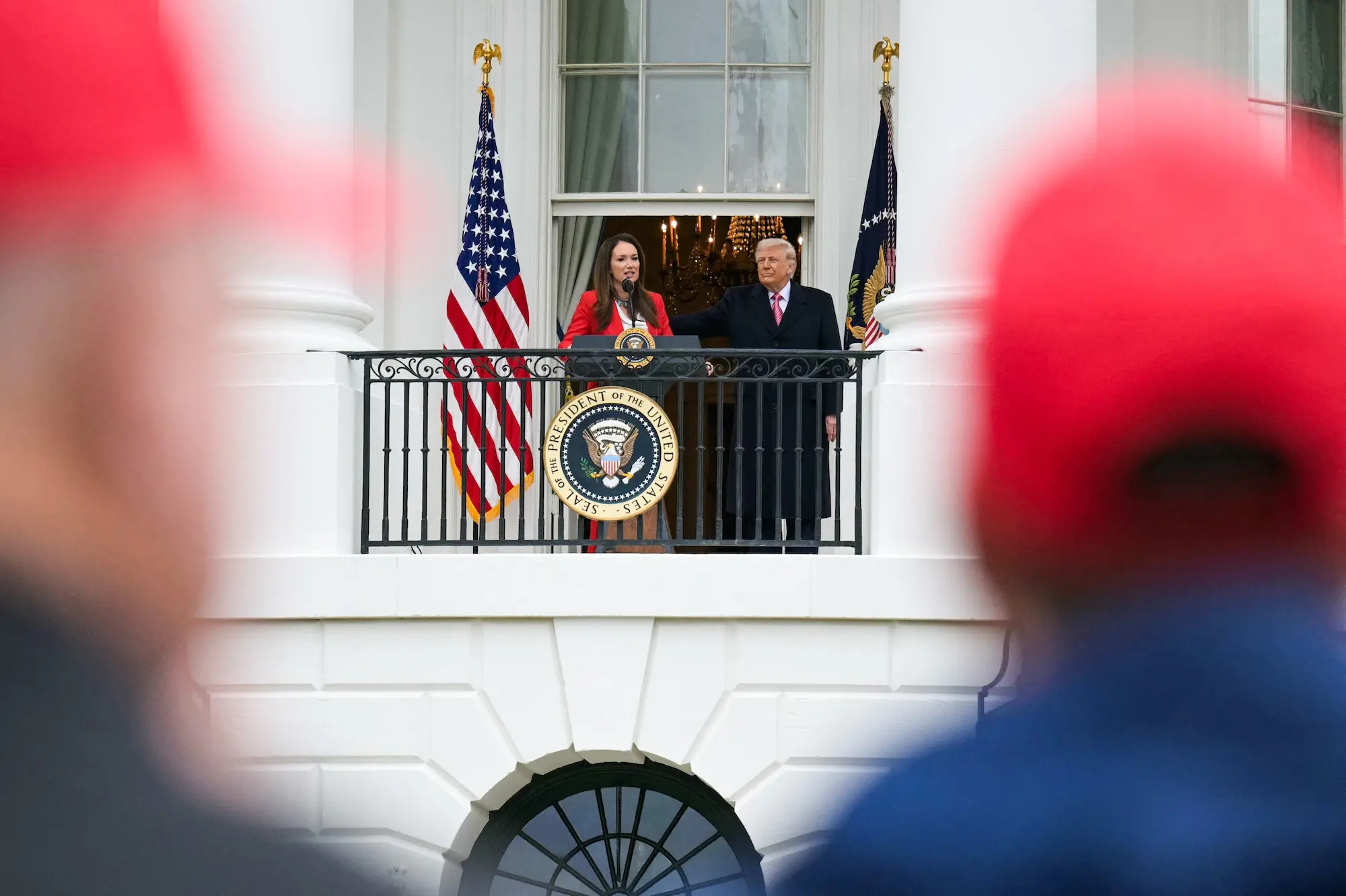 trump and rollins stand on a balcony with the presidential seal on it, as seen from among a crowd of people wearing red hats