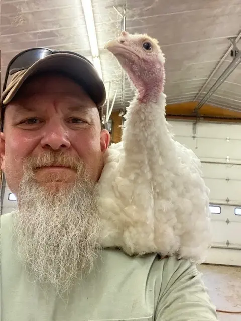 A man with a white beard poses for a selfie-style photo with a turkey or other bird sitting on his shoulder